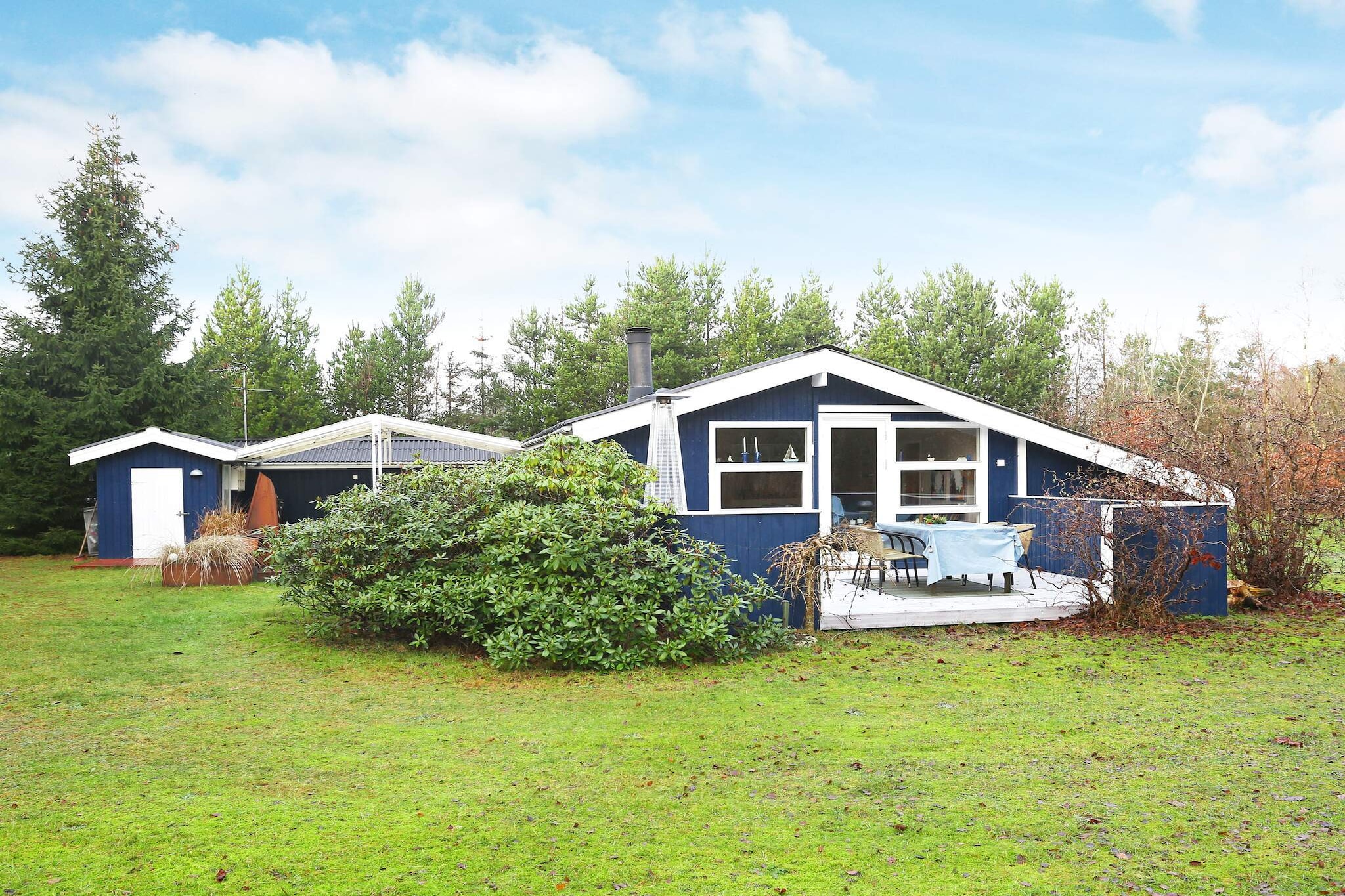 This vacation home is located in the attractive vacation area of Bratten, between Skagen and Frederikshavn, on a large and lovely natural plot with plenty of sunlight and lots of outdoor activities. The house is furnished with a well-equipped kitchen, a cozy living room with a wood-burning stove, a heat pump, and large windows that provide wonderful light. There are three good bedrooms with double beds and a bathroom with a shower.
The TV has built-in Chromecast, so you can stream your own services.
The cottage is located on a beautiful natural plot surrounded by trees, ensuring privacy. The living room opens onto a lovely south-facing terrace with a gas heater. There is also a covered terrace between the house and the shed, which also has an electric heater, so you have every opportunity to enjoy the Danish summer with sunbathing, playing on the grounds, and delicious barbecues. It is possible to spend the night in the shelter and cook at the campfire site. For the little ones, there is a play tower with a slide, swings, and a trampoline (use at your own risk/insurance). Lounge area with sleeping facilities. The owner provides 3 bicycles for adults (DanCenter does not guarantee their condition).
Approx. 400 m to a large communal playground with in-ground trampolines, large swings, and a nice soccer field. The beach is approx. 400 m away and offers white sand, calm water, a bathing jetty with picnic facilities, and more. In the summer, there is a grocery store and ice cream shop approx. 400 m away by the beach.