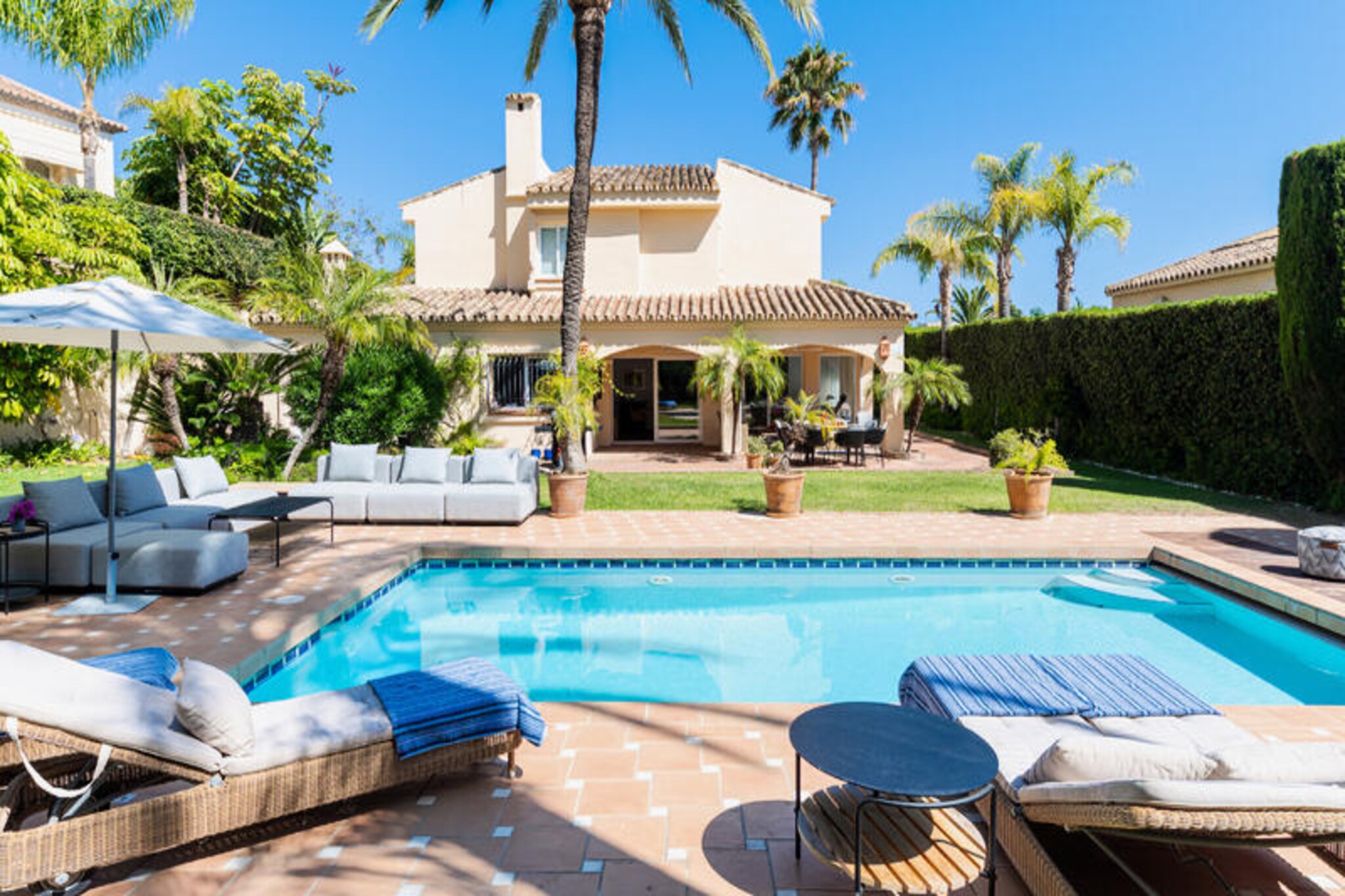 In the heart of the prestigious Los Monteros development, this exquisite Andalusian-style villa offers a serene retreat just moments from Marbella’s golden beaches and the exclusive La Cabane Beach Resort. Surrounded by lush gardens and the gentle Mediterranean breeze, this property invites you to experience the perfect blend of luxury, comfort, and Andalusian charm. Whether you’re seeking a tranquil escape or an active holiday, this villa’s prime location ensures you’re close to Marbella’s vibrant center, a local supermarket, the Los Monteros Racket Club, and the area’s top attractions.
Step through the antique wooden door into a world of elegance. The villa’s layout is thoughtfully designed, with four beautifully appointed bedrooms on the first floor. The master bedroom boasts an en-suite bathroom and a private balcony with a cozy seating area, ideal for soaking in the views. A children’s bedroom with twin beds and a single bedroom share a bathroom, while an additional double bedroom features its own en-suite. On the ground floor, a charming bedroom with a double bed and en-suite bathroom opens directly onto the terrace and garden. The spacious living room, complete with plush sofas and a large flat-screen TV, flows seamlessly into the dining area and a fully equipped blue kitchen, perfect for creating memorable meals. The covered terrace, with its inviting sofa and dining table, is the perfect spot for al fresco dining, complete with a barbecue for summer evenings.
The villa’s outdoor spaces are equally captivating. A large swimming pool, surrounded by sun loungers, an outdoor sofa, and beanbags, provides the ideal setting for relaxation under the Andalusian sun. The lush garden offers privacy and tranquility, making it a haven for families or groups of friends.
Whether you’re exploring Marbella’s vibrant nightlife, enjoying a game of tennis at the nearby racket club, or simply unwinding by the pool, this villa promises an unforgettable stay. Discover the essence of Andalusian elegance and create lasting memories in this exceptional Los Monteros retreat.